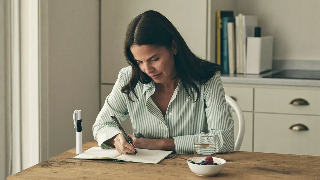 Woman writing in a symptom journal at her kitchen table next to an injection pen and a glass of water