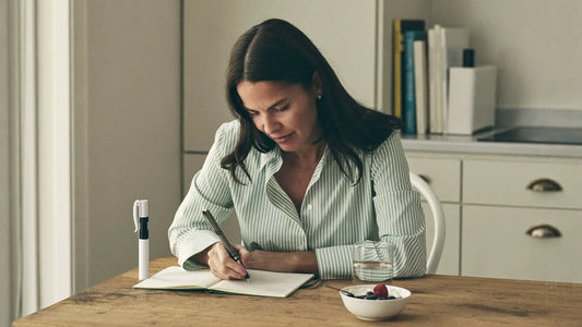 Woman writing in a symptom journal at her kitchen table next to an injection pen and a glass of water