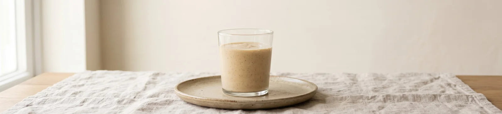 A glass of banana protein smoothie on a small plate on a linen runner