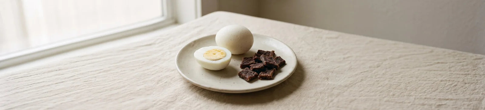 A whole hard-boiled egg, a halved egg and pieces of beef jerky on a small cream plate