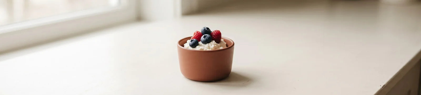 A small terracotta cup of ricotta topped with raspberries and blueberries on a light counter