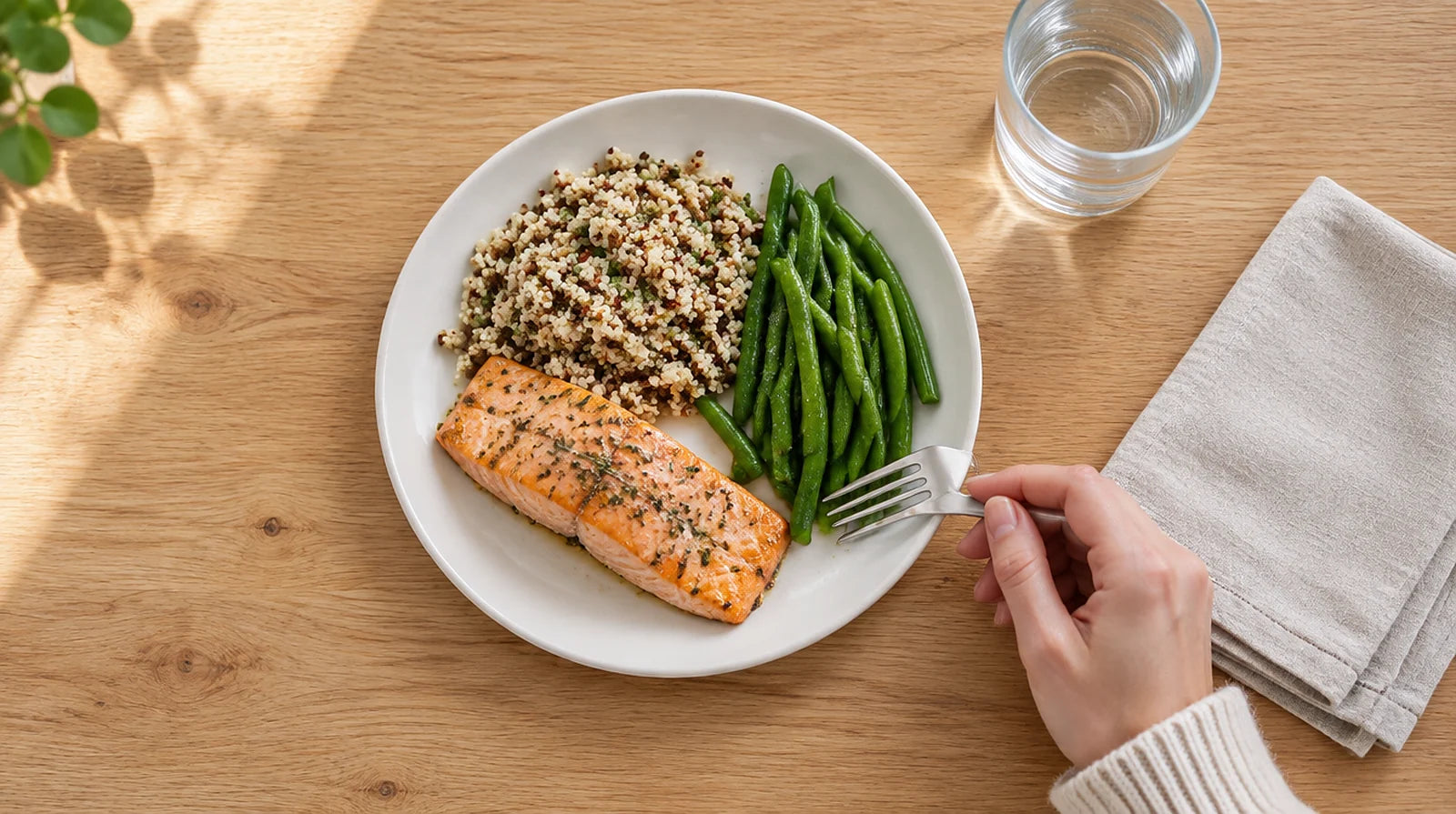 Overhead view of a small plate of baked salmon, quinoa, and green beans beside a glass of water and a linen napkin, with a woman's hand holding a fork